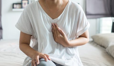Woman sits on the edge of her bed holding her chest in heartburn pain.